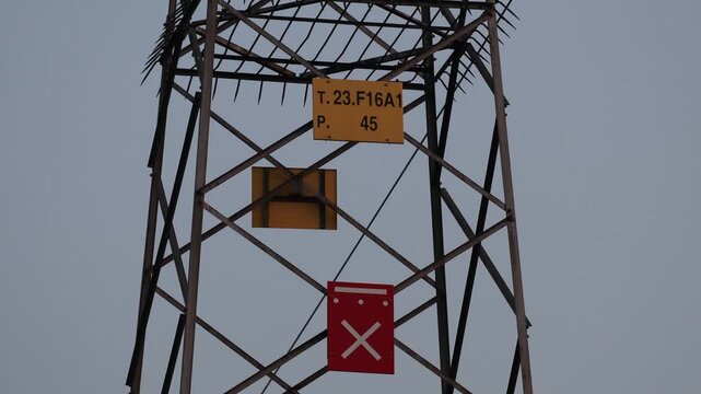 Close-up shot of the steel lattice structure of a high voltage power pylon featuring identification and warning signs against a plain sky background.