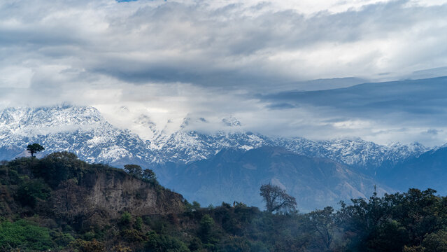 Snow-Capped Mountain Range Beneath Dramatic Clouds and Forested Hills
