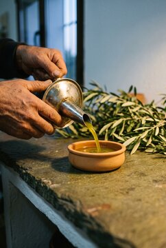 Hands pouring fresh extra virgin olive oil from a traditional metal dispenser into a terracotta bowl with olive branches in the background