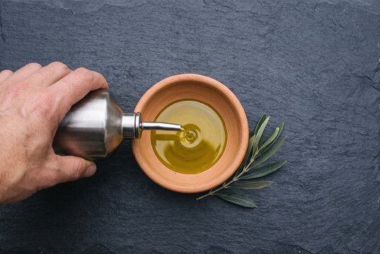 Hand pouring extra virgin olive oil from a metal dispenser into a terracotta bowl with an olive branch on a dark slate background