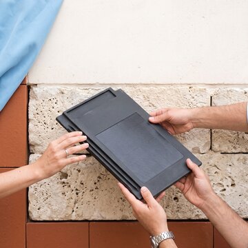 Hands holding a modern solar roof tile with integrated photovoltaic panel against a textured stone wall background