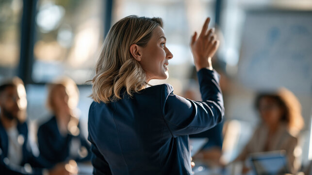 Confident businesswoman photographed from behind while presenting ideas to attentive colleagues in bright modern office professional attire raised hand gesturing toward