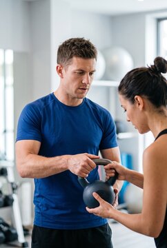 Male personal trainer demonstrating proper kettlebell holding technique to a young woman during a workout session in a gym