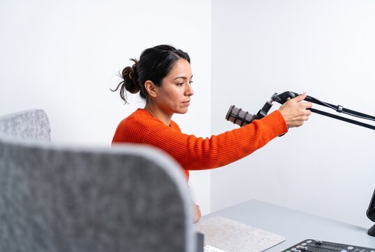 Hispanic woman adjusting a professional microphone while sitting at a recording studio desk for podcasting or broadcasting