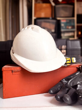 White hard hat resting on a red metal toolbox, with work gloves and a measuring tape, depicting labor, construction, and safety for manual workers and skilled trades
