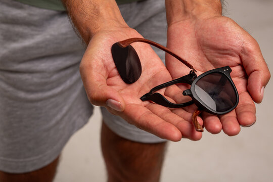A young guy in a colored T-shirt and gray shorts holds broken glasses in his hands after unsuccessfully transporting luggage at the airport. The glasses are made of low-quality material