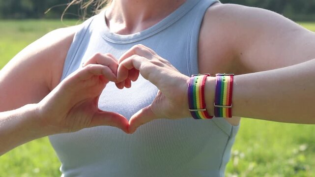 Female hands show heart gesture on blurred natural background. Woman with rainbow LGBTQ+ community bracelet on wrist. Girl makes heart shape with fingers, close-up. Love and tolerance concept