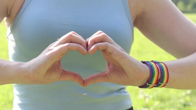 Female hands show heart gesture on blurred natural background. Woman with rainbow LGBTQ+ community bracelet on wrist. Girl makes heart shape with fingers, close-up. Love and tolerance concept