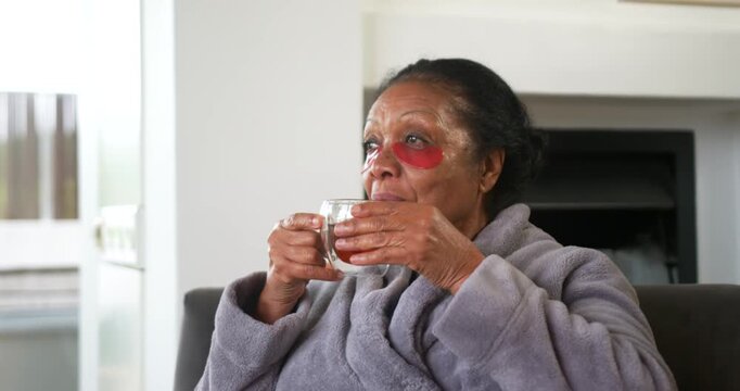 Senior African woman robe-clad lifting mug, sipping, turning to window with red eye patches resting