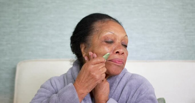 Senior African American in robe on sofa bringing gua sha to lips, gliding jawline for skincare
