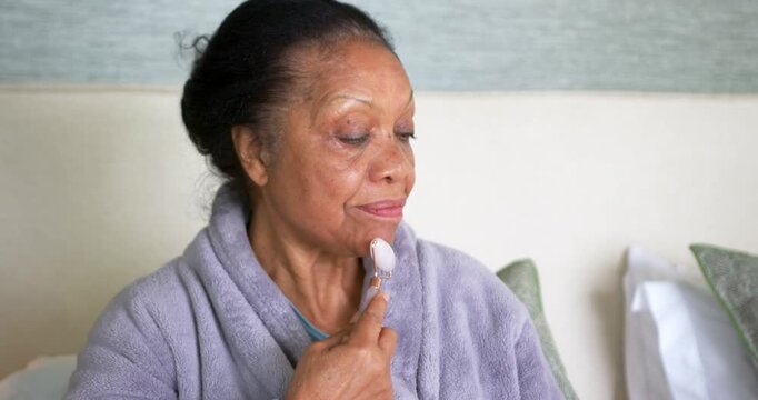 Senior African woman in robe on bed rolling rose-quartz roller upward jawline-to-eye for blood flow