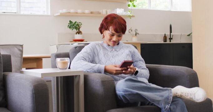 African American woman holding pink phone sitting at home in armchair scrolling and checking texts