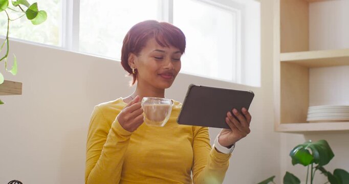 African American woman sitting by window in mustard top holding tablet laughing at clip sipping mug