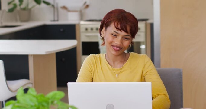 African American woman sitting in kitchen smiling leaning, reacting to laptop content with plant