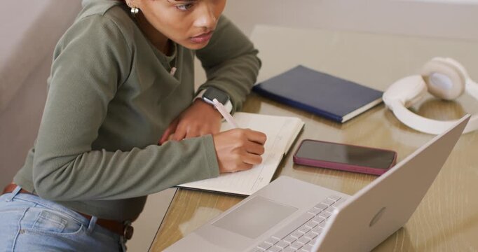 African American woman leaning on notebook, taking notes prompted by laptop and phone in olive top