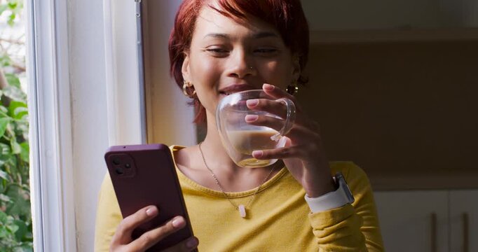 20s woman reading purple phone after message, laughing, sipping double-walled mug at kitchen window