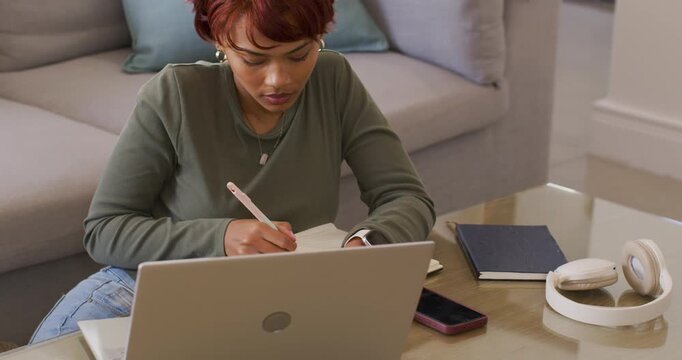 African American woman transcribing from laptop, writing notes on notebook with pink pen at home