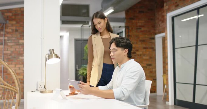 Diverse coworkers in office clothes, woman approaching, man tapping tablet at desk showing content
