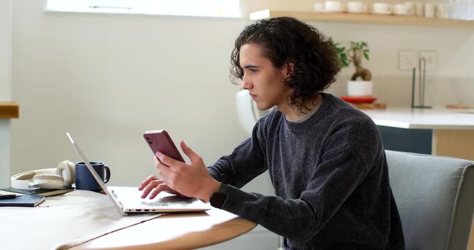 African American man 20s at kitchen table, glancing at pink phone, typing on laptop for work