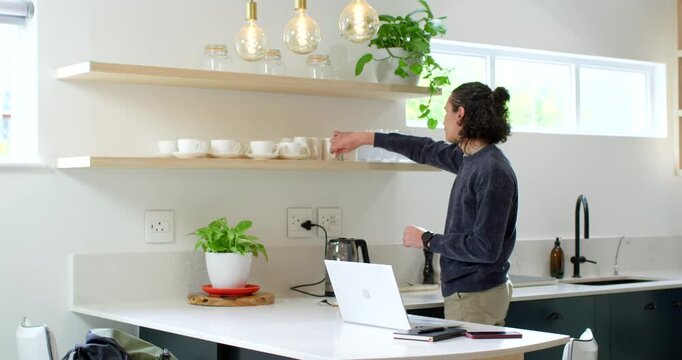 Man in 20s at kitchen island noticing cups on shelf, reaching cup, toggling kettle, preparing drink