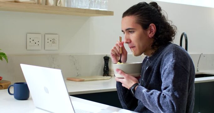 African American man in sweater working on laptop, eating bowl with spoon and typing at counter