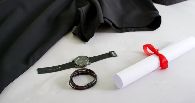 Camera panning zooming revealing black gown, watch, bracelet, diploma on white fabric for grad prep