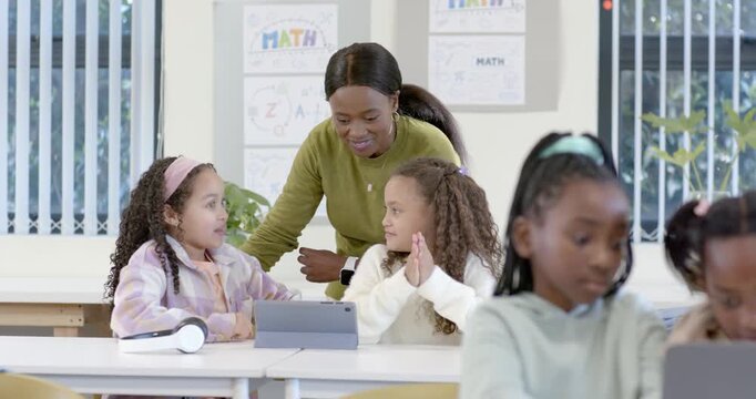 African American girls presenting tablet in class, teacher leaning and gesturing, showing learning