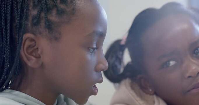 African schoolgirls speaking at desk, left girl whispering secret, right girl reacting by laptop