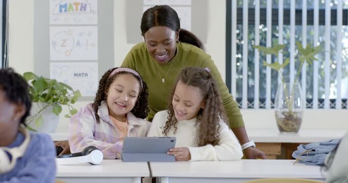 Teacher with two female teens leaning over desk one pointing tablet, reacting to clip and laughing