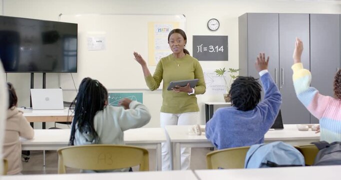 African American teacher asking question with tablet in class, female teens raising hands answering