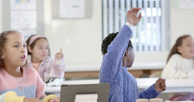 School-age girls during lesson, blue-sweater girl raising then reaching then resting by tablet