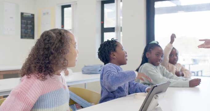 African American girls raising hands and talking in class as teacher coming cupping ear with tablet