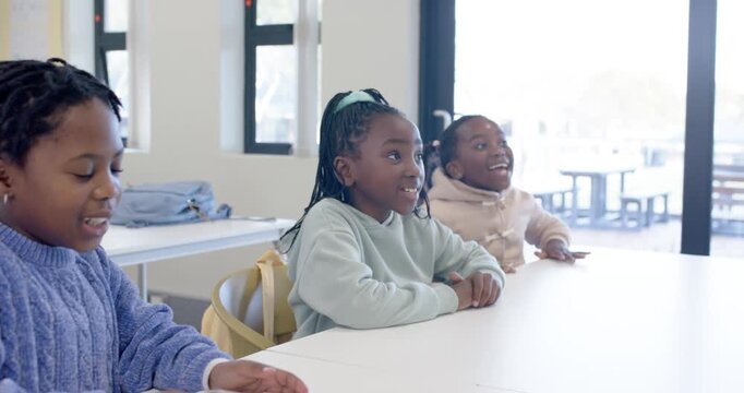 African American schoolgirls watching, reacting to off-camera speaker at long white table in class