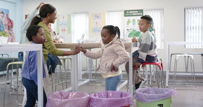 African American teacher holding bottle, girl in pink coat sorting into red GLASS bin for recycling