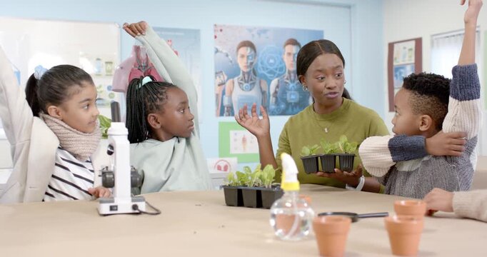 African American teacher holding seedling tray explaining plant growth, kids raising hands at table