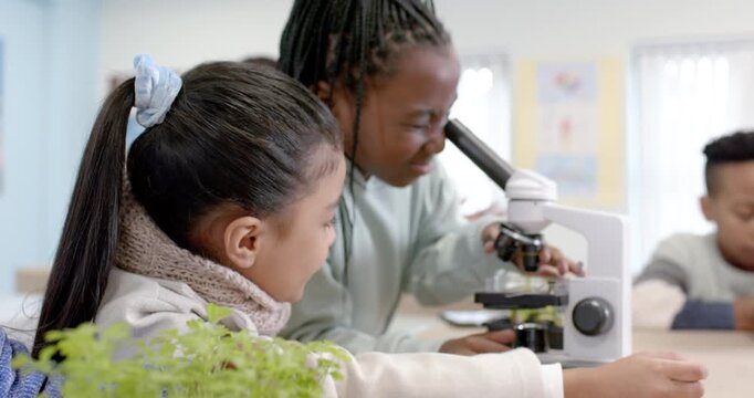 African American class watching braided girl operating microscope with teacher guiding near plants