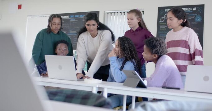 Indian instructor leaning and pointing at laptop at table, guiding teens using tablet and AI poster
