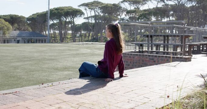 Teenage female in maroon hoodie sitting on brick ledge by turf, noticing camera turning and smiling