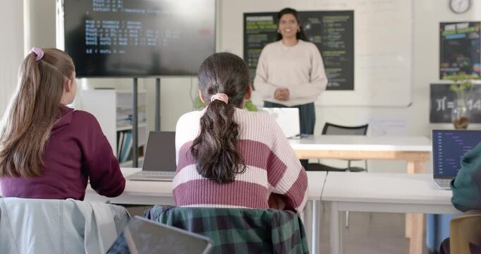 Indian teacher leading class, speaking and pointing to screen and laptops, teaching teens typing