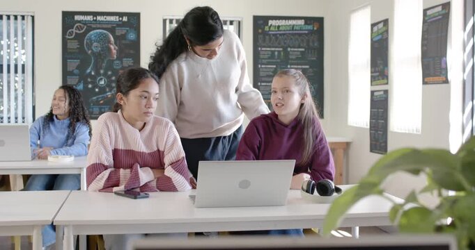 Indian tutor leaning, pointing to silver laptop helping youth females fixing code on monitor in lab