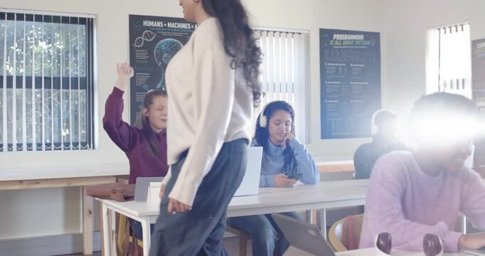 Indian teacher walking in class toward teen raising hand leaning pointing at laptop as peers using