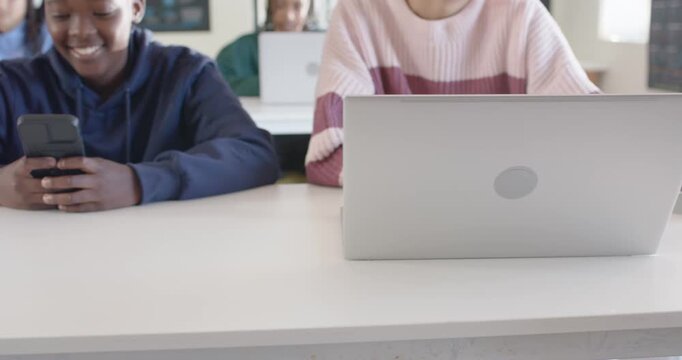 Teen in hoodie typing, smiling on phone in class as camera pulls back, glancing at classmate