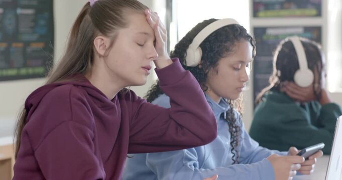 Three youths sitting in class, middle student using phone with headphones, causing left fidgeting
