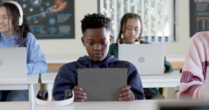 Teen classmates working in class, navy-hoodie teen holding tablet, others typing laptops