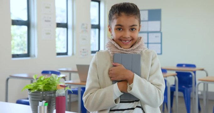 Camera filming teen girl wearing beige coat, scarf standing front class hugging tablet, smiling