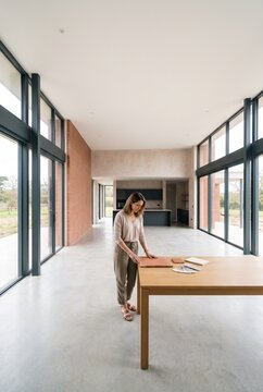 Female interior designer inspecting tile samples and material swatches on a wooden table in a spacious modern architectural house with large windows