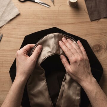 Hands of an artisan seamstress carefully adjusting the lining of a dark coat collar on a wooden workbench surrounded by sewing tools