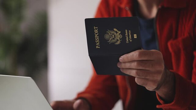 Young male holding American passport while applying for a visa on laptop