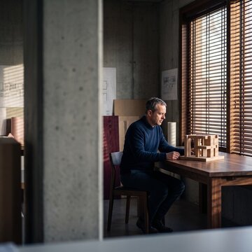 Focused male architect sitting at a wooden desk examining a miniature scale architectural model in a modern concrete studio