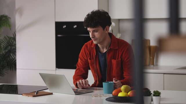 Young male typing on laptop and drinking tea at home, communicating online